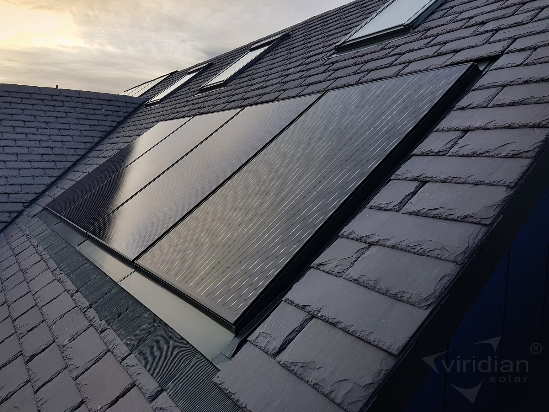 Rooftop solar panels installed on a slate-tiled roof under a cloudy sky.