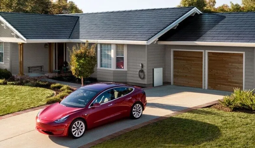 A red car is parked in a driveway in front of a single-story house with solar panels on the roof. The garage has wooden doors, and the lawn is neatly maintained.