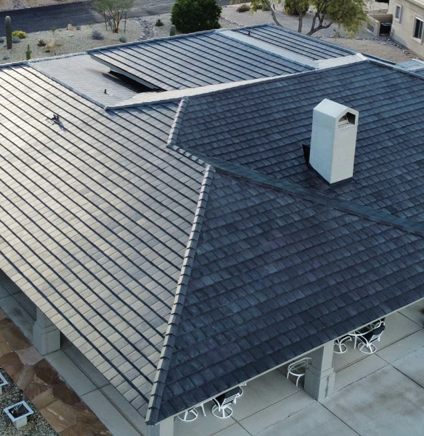 Aerial view of a house with a dark gray, shingled roof, featuring a skylight and a chimney. The surrounding area includes a landscaped yard with desert plants and outdoor seating.