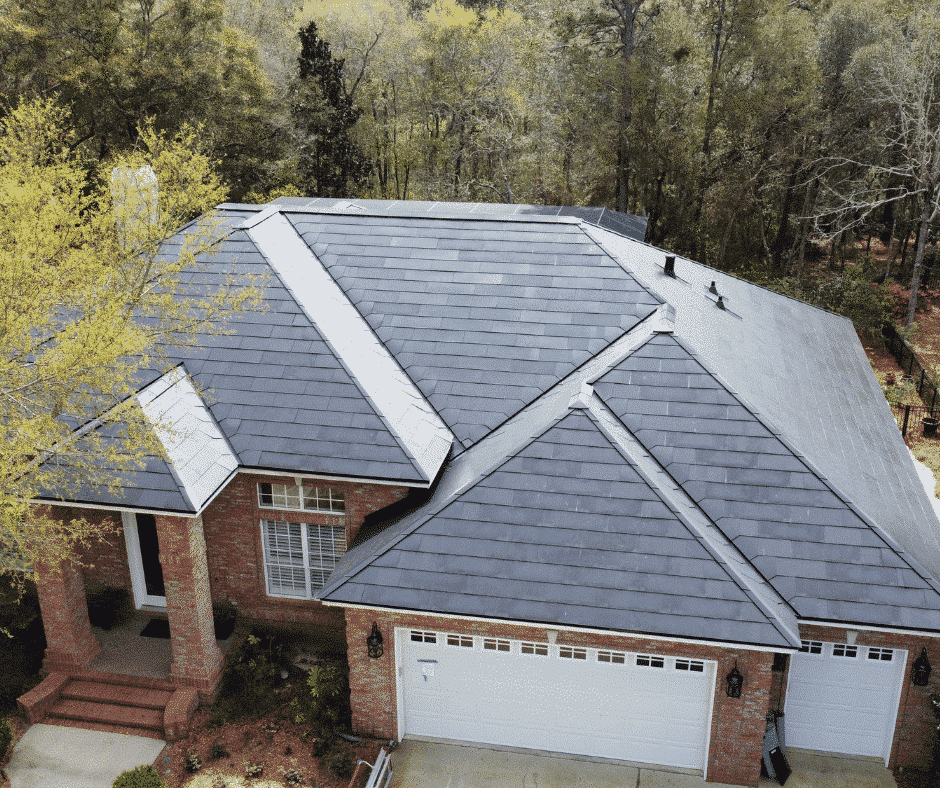 Aerial view of a house with a sleek, dark-colored metal roof surrounded by trees. The house features a three-car garage and a brick exterior.