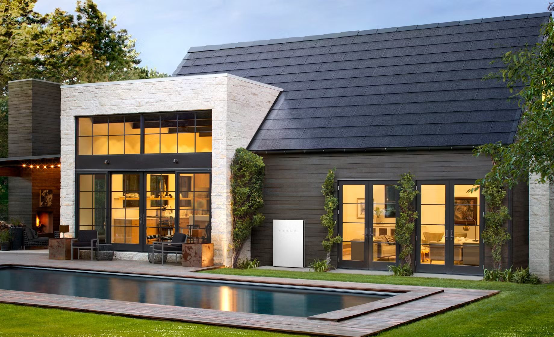 Modern house with large windows, solar roof tiles, and a rectangular pool in the foreground. Lawn surrounds the pool, and the house features multiple stories with evening lights on inside.